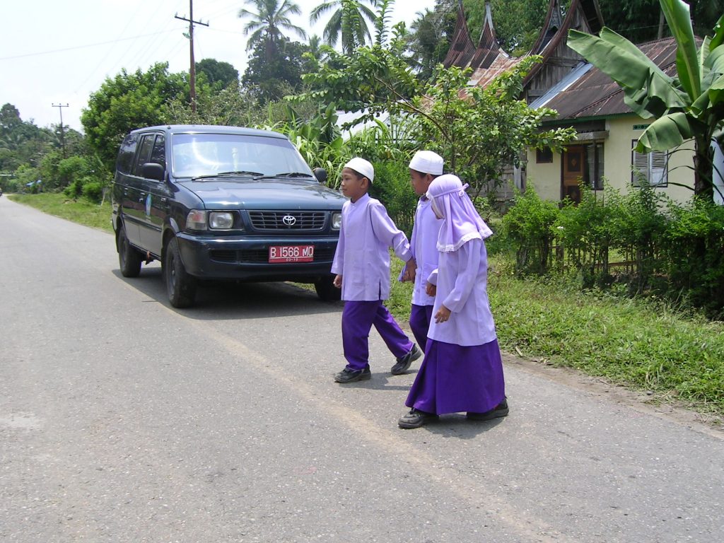 Two boys and a girl are holding hands. They are walking across a road. A car is approaching and is very close to them.