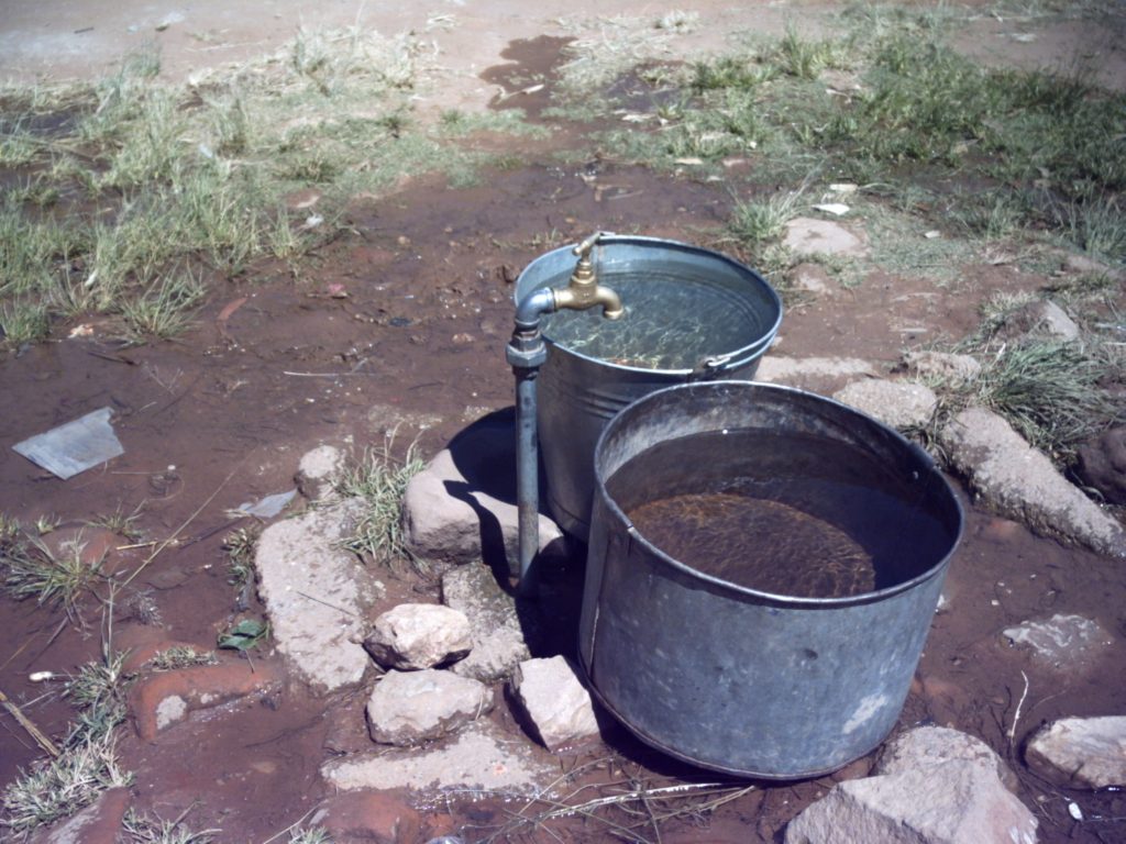 Two metal buckets filled with water stand under a tap. The ground around the tap is earth with many large stones