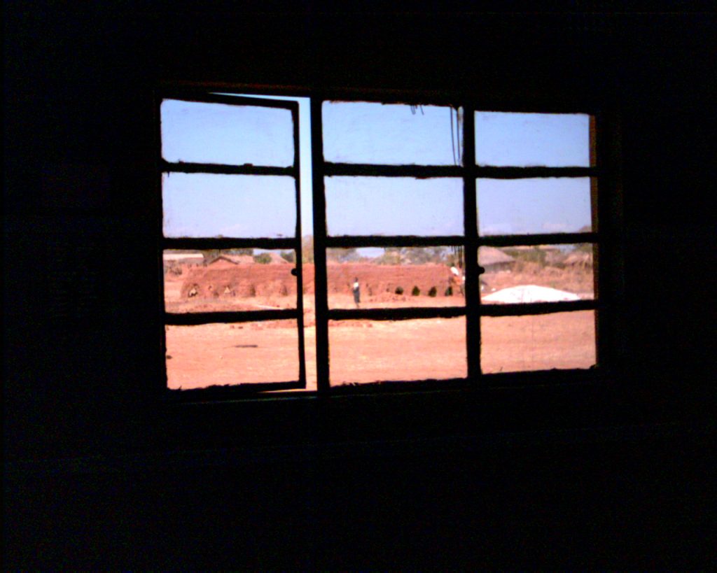 The picture was taken from inside the classroom, looking out of a window. The inside of the room is very dark so no details are visible. Outside the window there is blue sky, bare earth and in the background a huge pile of bricks