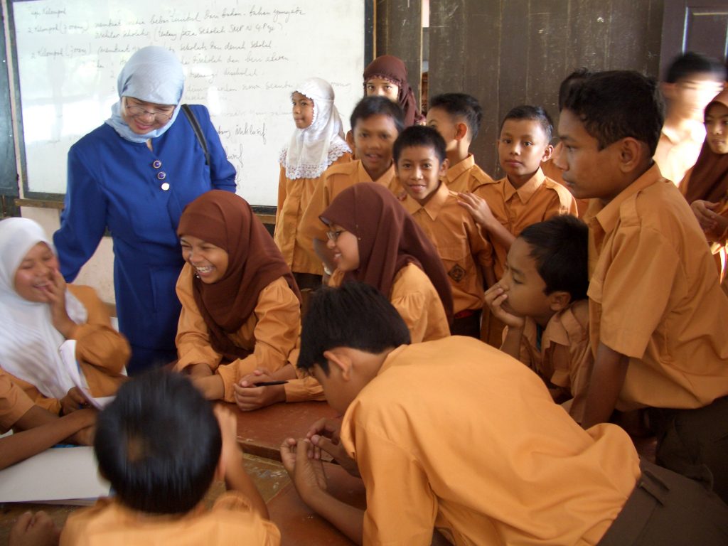 Girls and boys are sitting around a table, with other children standing behind them. A smiling female teacher is talking to some of the girls who are laughing. The boys are listening/watching but not laughing.]