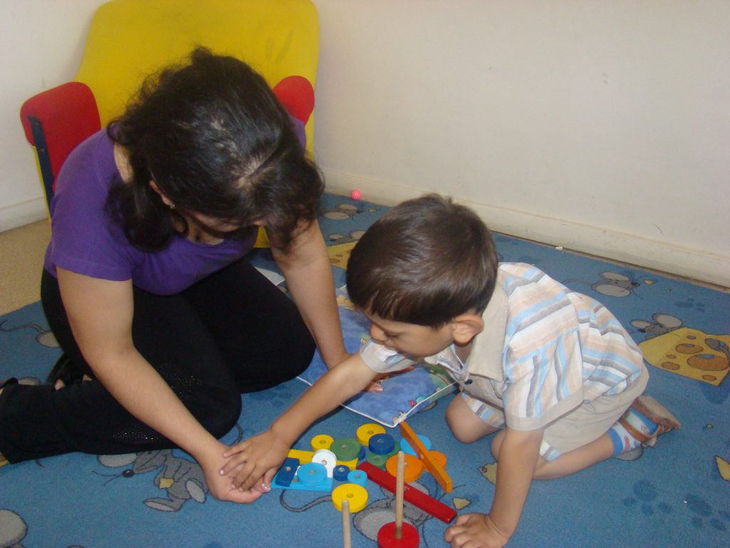 A woman and a young boy are kneeling on the floor. They are playing with a toy that involves putting disks of plastic onto wooden sticks